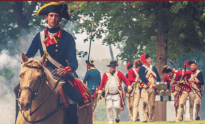 A man in 18th-century military uniform rides a tan horse in the foreground, while several soldiers in red coats march behind him during the Ticonderoga StreetFest in a wooded outdoor setting.